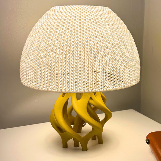 A table lamp with a large white textured dome shade and a sculptural yellow base, shown on a white tabletop against a gray wall.