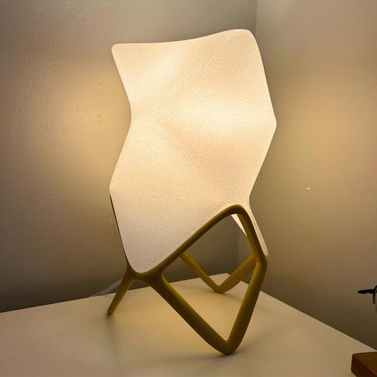 A modern sculptural table lamp with a curved, wave-shaped textured white shade and a geometric diamond-shaped gold base, shown lit against a neutral wall.