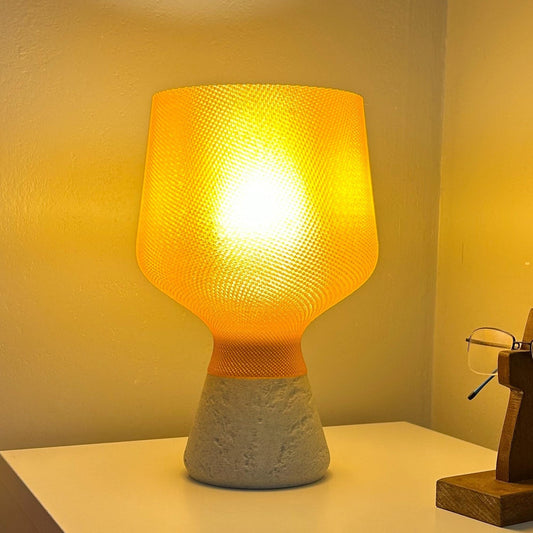 A table lamp with a glowing yellow-orange textured shade and a textured gray concrete base, sitting on a white surface against a warm beige wall.