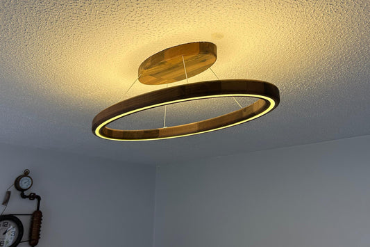 A modern double-ring LED ceiling light fixture with a warm wood finish, suspended from the ceiling in a room with a textured white ceiling and light gray walls.