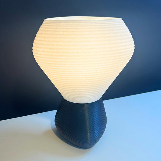 A modern table lamp with a ribbed, bulb-shaped white shade and a dark navy blue textured base, shown lit against a dark blue background.