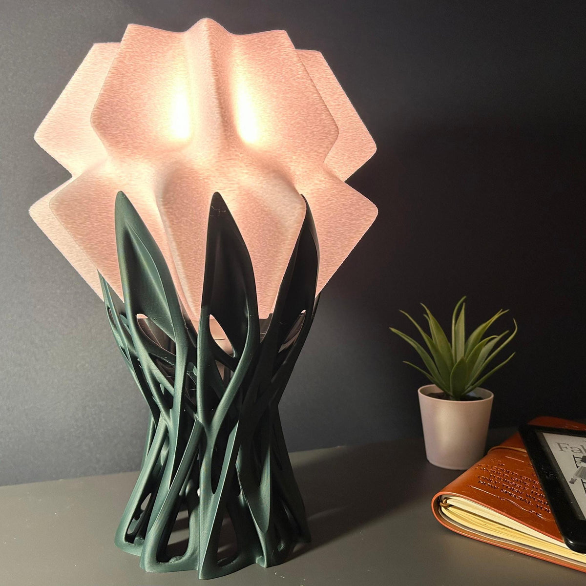 A sculptural table lamp with a glowing pale pink faceted shade and a dark green twisted base, shown on a tabletop beside a small potted plant and an open notebook against a dark gray wall.