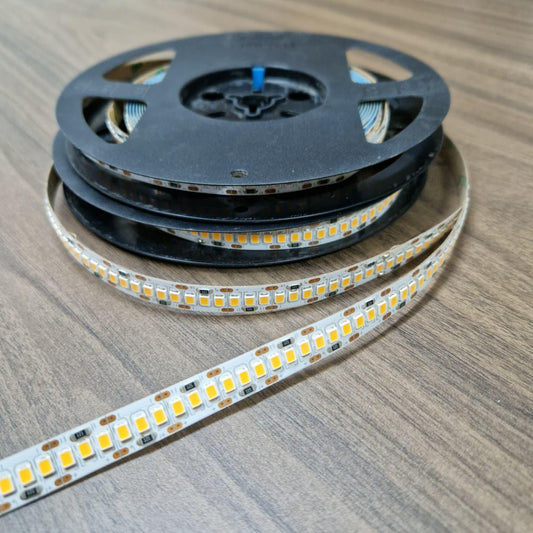 A spool of LED strip light with several coils wound around a black plastic core, with one coil unrolled to show the flexible strip. The strip features small yellow LED chips on a silver base with a white protective tape, set against a brown wood-grain tabletop.
