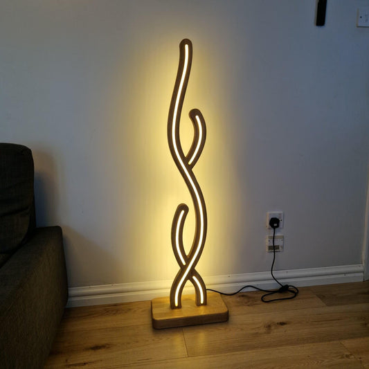 A modern abstract floor lamp with a glowing vertical S-shaped design on a square wooden base, placed on a wooden floor against a light gray wall.