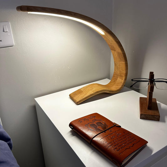 A curved wooden LED desk lamp with a warm glowing light, displayed on a white side table next to a brown leather journal and a wooden eyeglasses stand.