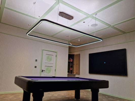 A modern rectangular LED light fixture with a black frame hangs from the ceiling of a room with a purple pool table in the foreground, ribbed cream walls, and a large black wall-mounted screen.