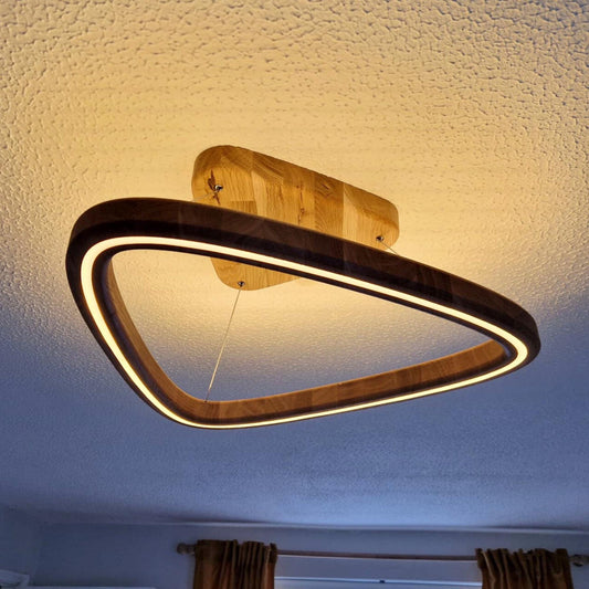 A modern triangular LED light fixture with a dark wood frame and a warm glowing light, mounted on a textured ceiling in a room.