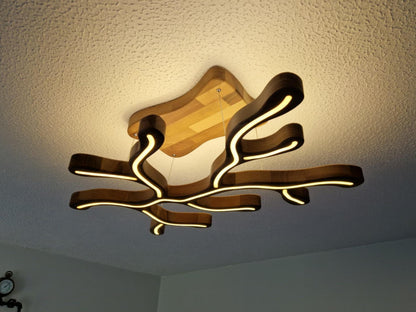 An abstract, organic-shaped ceiling light fixture made of layered dark wood and illuminated with warm white LED strips, shown installed on a textured white ceiling.
