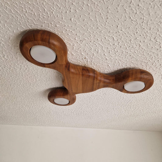 A wooden flush mount ceiling light with a sculptural, organic three-armed shape and three white circular light covers, mounted on a textured white ceiling.
